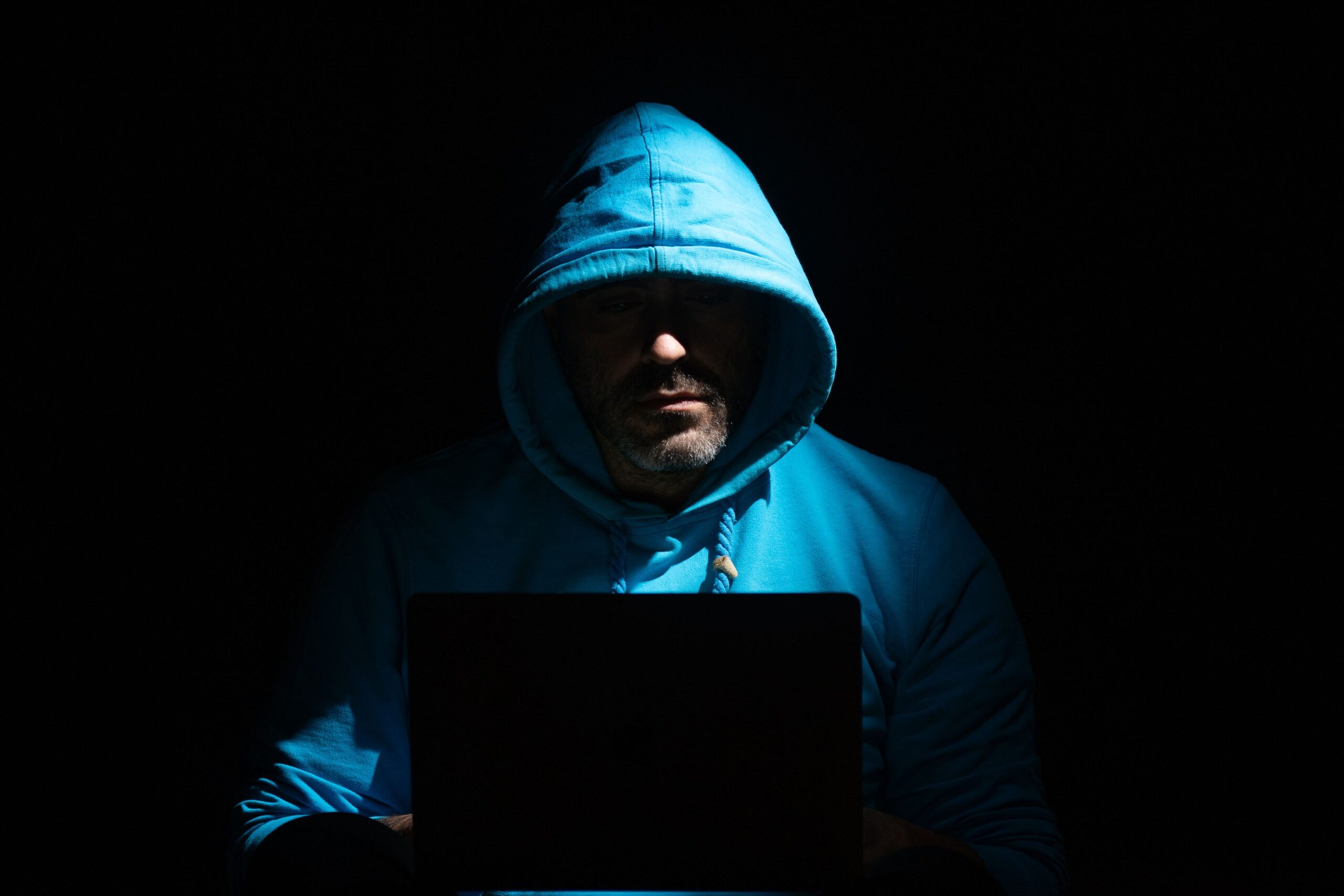 Privacy Policy A man in a blue hoodie focused on a laptop in a dark room, conveying secrecy and intrigue.